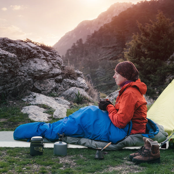 Camper in a red jacket sitting in a blue sleeping bag beside a yellow tent, enjoying a hot drink in a mountainous outdoor setting at sunrise.